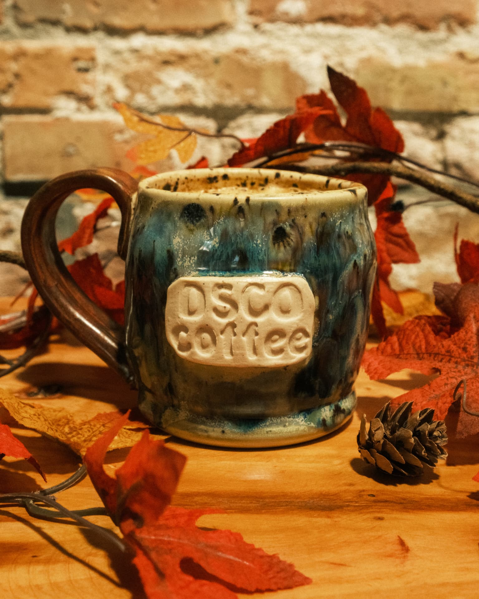 A handmade DSCO Coffee mug surrounded by autumn leaves and a pinecone